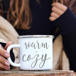 Load image into Gallery viewer, Picture of 11 oz White Enamel Camp Coffee Mug with Silver Rim.  Graphic of Warm and Cozy in Grey colored font. Woman is cozied in wrap.
