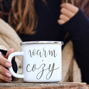 Picture of 11 oz White Enamel Camp Coffee Mug with Silver Rim.  Graphic of Warm and Cozy in Grey colored font. Woman is cozied in wrap.