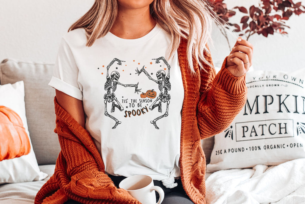 Picture of White Hallloween T-Shirt with black dancing skeletons, pumpkins and stars.  The graphic says, Tis the season to be spooky