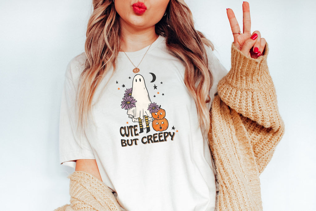 Picture of woman in Halloween T-Shirt, Cute But Creepy in Black letters, Girl with sheet for Halloween Costume holding purple daisies. Two orange pumpkins look on.