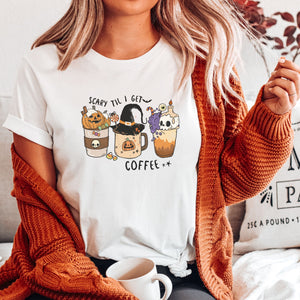 Picture of young woman in burnt orange colored cardigan wearing our Soft cream women's t-shirt for halloween with graphic of three coffee drinks, one with a pumpkin, the center with a witches hat and the last with a skull candle.